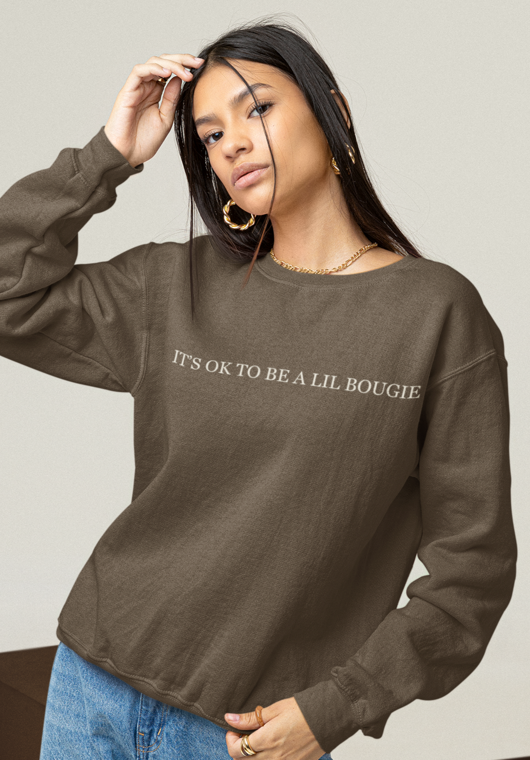 Model wearing brown Bougie crewneck with “It’s OK to Be a Lil Bougie” printed across front. Brushing hair out of face