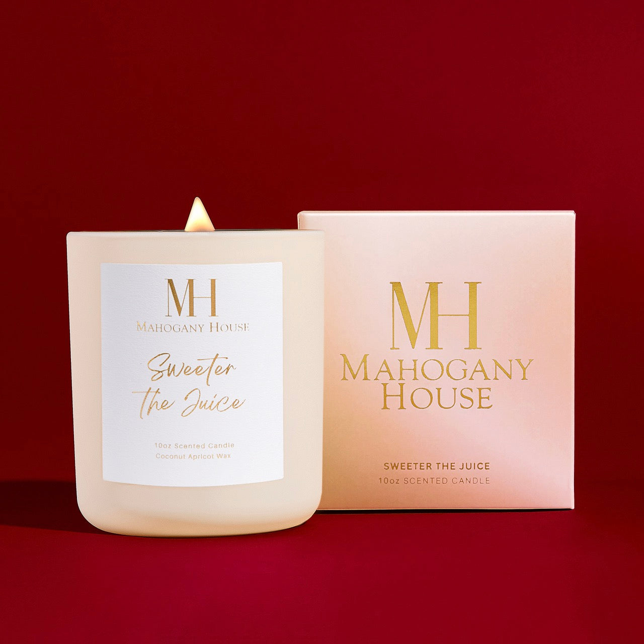 Lit Sweeter The Juice wooden wick candle in white glass vessel beside Mahogany House blush pink packaging, styled on a red background.