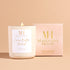 Lit Cocoa Butter Kissed 10oz wooden wick candle in white glass vessel with gold lid, placed beside its blush pink Mahogany House box on a neutral background.