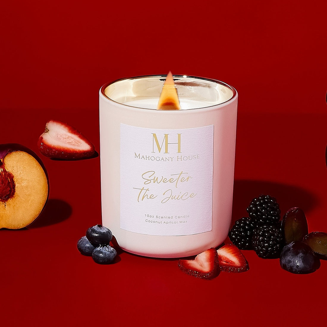 Lit Sweeter The Juice 10oz wooden wick candle in white glass vessel, styled on a red background with blackberries, strawberries, blueberries, grapes, and sliced plum.