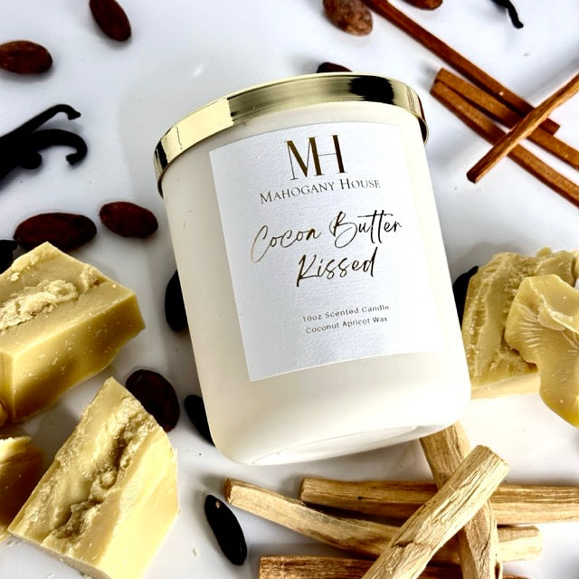 Unlit Cocoa Butter Kissed wooden wick candle in white glass vessel with gold lid, laying flat surrounded by cocoa butter chunks, vanilla sticks, and cocoa beans on a bright white background.