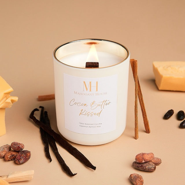 Lit Cocoa Butter Kissed 10oz wooden wick candle in white glass jar with gold lid, styled with vanilla beans, cocoa pieces, and spice accents on a warm-toned surface.