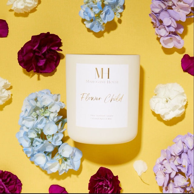 Unlit Flower Child 10oz wooden wick candle in cream glass vessel, styled on a yellow background with purple, blue, and white hydrangea flowers.