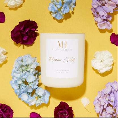 Unlit Flower Child 10oz wooden wick candle in cream glass vessel, styled on a yellow background with purple, blue, and white hydrangea flowers.