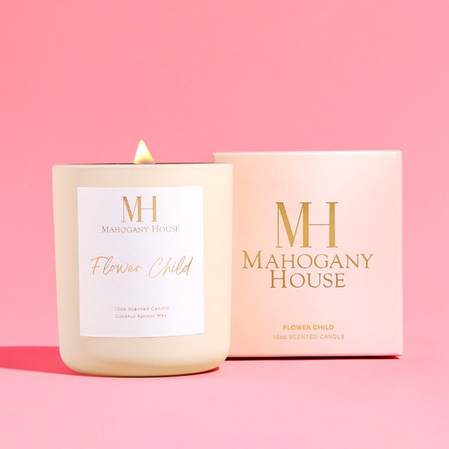 Lit Flower Child 10oz wooden wick candle in cream glass vessel with gold lid, displayed beside blush Mahogany House packaging on a pink background.