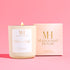 Lit Flower Child 10oz wooden wick candle in cream glass vessel with gold lid, displayed beside blush Mahogany House packaging on a pink background.