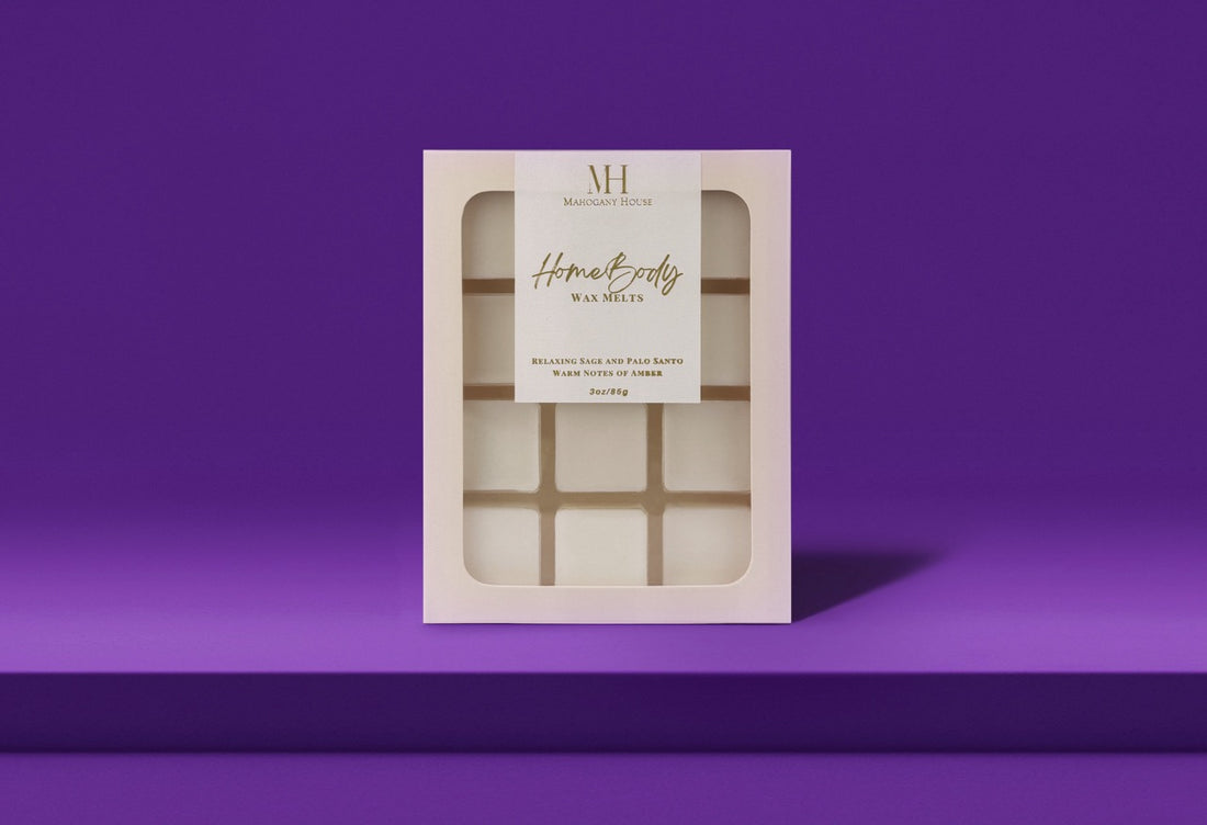 HomeBody wax melts in white clamshell packaging with Mahogany House branding, styled with palo santo, sage bundles, and blackberries.