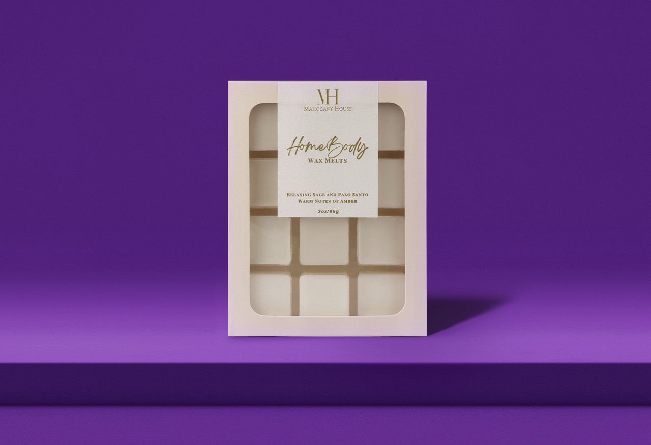HomeBody wax melts in white clamshell packaging with Mahogany House branding, styled with palo santo, sage bundles, and blackberries.